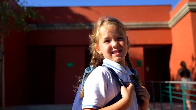 Beautiful elementary school girl with pigtails and a backpack smiling and waving goodbye to her mother before entering the school building on a sunny day, ready for a new day of learning