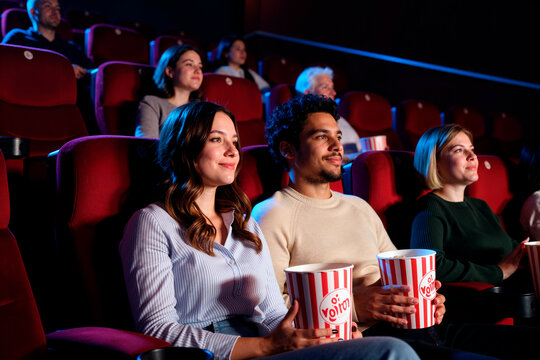 Young adult Caucasian woman and young adult multiethnic man sitting in movie theater holding popcorn, watching screen with other audience members of various ages in background - Powered by Adobe