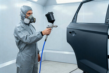 Professional auto body painter in protective suit and respirator applying primer or paint with a spray gun to a car door panel inside a specialized paint booth.