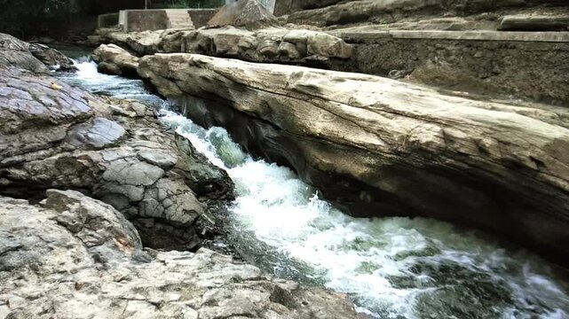 The video shows a gently flowing river passing through the Lava Bantal area in Berbah, Yogyakarta. The water moves steadily between dark, rounded basaltic rocks that line both sides of the river.