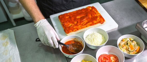 Banner of male chef hand with gloves scooping tomato sauce from bowl while preparing focaccia on stainless steel counter, surrounded by fresh topping like cheese, tomatoes, corn and mixed vegetables