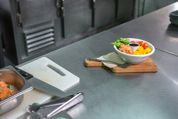 Salmon poke bowl with edamame, mango, and cherry tomatoes served over wooden board on a stainless steel counter of empty professional kitchen, focusing on healthy eating and modern cuisine preparation