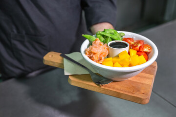 Close up of chef hand showing freshly gourmet poke bowl, featuring salmon, edamame, mango and tomatoes, served over wooden board in a professional kitchen environment. Healthy and tasty food concept.