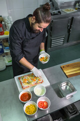 Male chef with apron and gloves in a professional kitchen preparing a delicious vegetarian focaccia, surrounded by bowls of fresh cheese, corn, tomatoes and mixed vegetables.