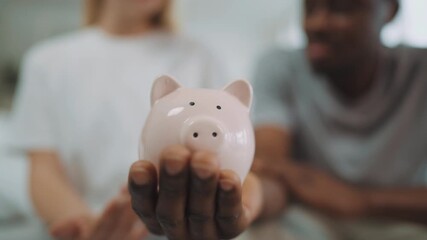 Closeup of funny ceramic piggy bank in hand of black man, woman putting coins. People save money for buying house and dreams realizing, family budget and finance, happy multiracial married couple - Powered by Adobe