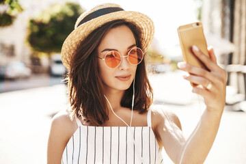 Teen female in summer white hipster clothes in headphones taking selfie on smartphone on warm sunset background