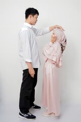 A photo of a tall Asian man gently touching the head of a shorter woman in a hijab, both smiling against a white background.
