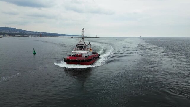 Aerial view of a red tugboat sailing through the port channel toward open sea. Maritime navigation and harbor operations concept