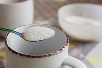 Adding granulated sugar into cup, closeup photo. Adding sugar to a mug with rainbow spoon on breakfast table, close-up, rustic style