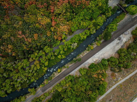 Aerial view of a dark river snaking through a vibrant tapestry of autumn foliage, juxtaposed against pale pathways, American Fork, Utah, United States.