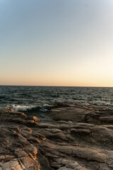 Waves softly lap against the rugged rocks along the shoreline as the sun sets, casting warm colors across the sky and reflecting on the water's surface.