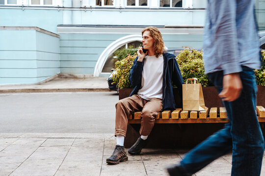 Young man sitting on bench while talking on phone in urban setting during the day