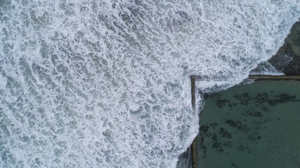 Aerial view of powerful waves crashing against the calm, rectangular ocean pool's edge, a stark contrast of textures and tones, Sydney, New South Wales, Australia.
