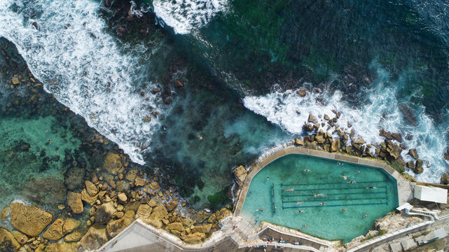 Aerial view of turquoise waves crashing against the rugged coastline and the calm, rectangular ocean pool filled with swimmers, Bondi Beach, Sydney, New South Wales, Australia.