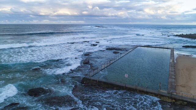 Aerial view of the ocean pool embraced by rocky shores and crashing waves under a cloudy sky at North Narrabeen, Sydney, New South Wales, Australia.