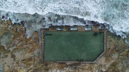 Aerial view of the turquoise ocean pool embraced by rugged, golden rocks, as frothy waves crash against the stone barrier, creating a mesmerizing contrast, Sydney, New South Wales, Australia.