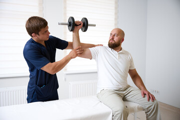 Physiotherapist assisting male patient with shoulder rehabilitation exercise using dumbbell. Man sitting on medical couch while performing physiotherapy workout under supervision of doctor.