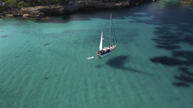 Aerial view of a sailboat gently floats on the clear turquoise waters near the rocky coastline, Balearic Islands, Spain.