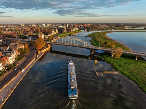 Aerial view of a cargo ship sailing under the Wilhelminabrug bridge reflecting the golden sunlight, contrasted by the autumn foliage along the IJssel river, Deventer, Overijssel, Netherlands.