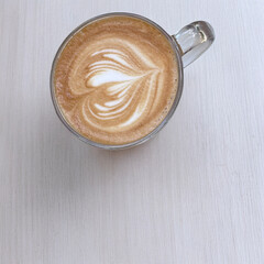 A glass mug of latte with a heart-shaped pattern of latte art sits on a simple white wooden table, viewed from above.
