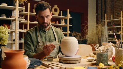 Caucasian sculptor man potter craftsperson ceramist small business owner male in apron carefully painting big ceramic vase jar paintbrush drawing design ornament pattern decorating pottery at workshop