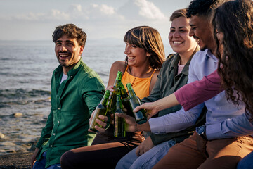 Group of diverse friends toasting with beer on the beach at sunset. Concept of gen z celebration, multi-ethnic friendship, summer vacation party, and enjoying alcohol together.