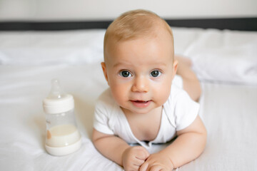 Beautiful smiling cute baby lying on stomach with a bottle of milk on bed. Children, people, infancy and age concept
