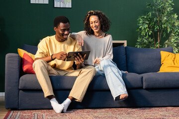Young diverse couple laughing sharing tablet on sofa