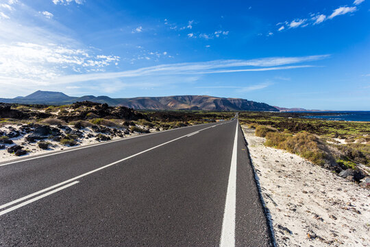 View of a seemingly endless road cutting through volcanic landscapes, under a vast blue sky with scattered clouds, leading to the sea, Lanzarote, Canarias, Spain.