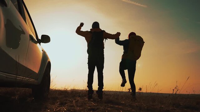 Couple walk from car at sunset silhouetted against sky on open field road holding hand and wearing backpack travel gear conveying travel and adventure with car silhouette and pair walking along road