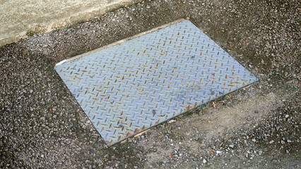 Metal Diamond Plate Access Cover on Rough Aggregate Ground: Industrial Texture, Urban Drainage, and Grunge Background