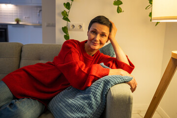 Obraz premium Smiling mature woman resting head on arm on sofa. Happy female looking camera on sofa, graceful retirement, natural wrinkles, confident female in 50s, mature happy aging, joyful relaxing at home