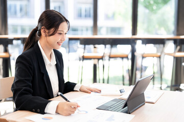 Fototapeta premium Asian businesswoman happily taking notes and reviewing financial data on paper and a tablet.