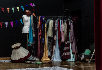 Backstage of a theatre with vintage costumes on a rack, a dress form, scattered props, and colorful bunting, capturing the behind-the-scenes atmosphere before a stage performance.