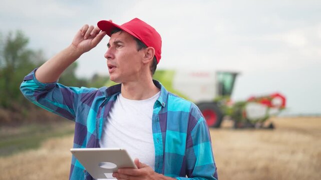 Farmer using tablet in field monitoring crop and combine harvester, showing agriculture technology and digital farm management with worker in cap and plaid shirt checking device data in rural