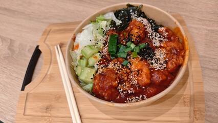 Korean rice bowl with spicy chicken, fresh vegetables, and sesame seeds served on a wooden tray