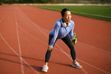 Obraz premium Female athlete resting on running track, breathing heavily, holding water bottle after exhausting exercise