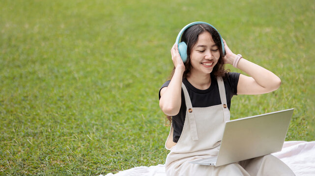 Happy college student enjoying music with headphones and laptop on campus lawn