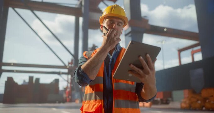  Male Logistics Worker in Safety Gear Uses a Tablet and Radio to Streamline Container Operations at a  Port, Optimizing Shipping Processes Amid Cranes and Cargo.