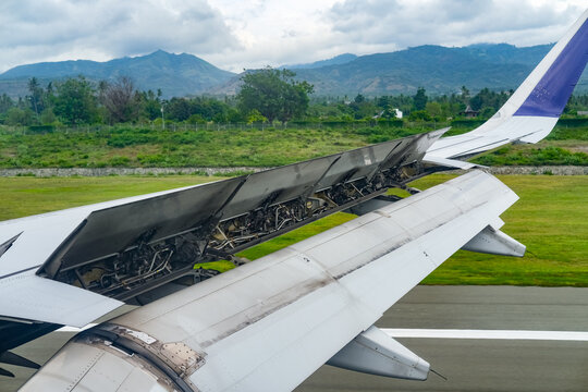 Aircraft wing with extended spoilers during landing rollout, showcasing aerodynamic braking in action as the plane touches down on the runway with green landscape and mountain views in the background.