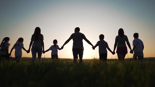 Holding hand of child in field at sunset, family silhouette crosses tall grass horizon, parent and child walking together in unity, community bond peaceful outdoor togetherness under warm sky