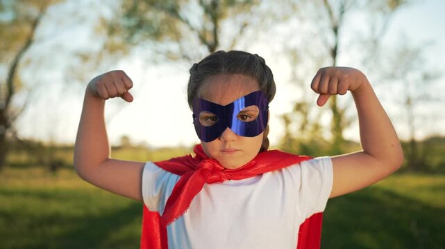 Child poses in superhero outfit flexing arm and showing strength while girl wears mask and red cape in outdoor park during play and roleplay portrait conveying confidence imagination courage and fun
