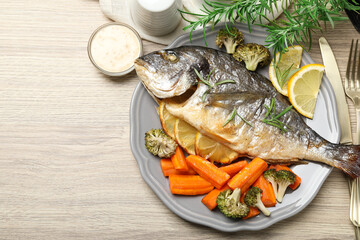 Cooked fish served with rosemary, lemon and vegetables on wooden table, flat lay. Space for text
