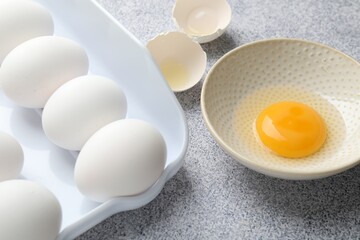 Many whole raw chicken eggs and broken one in bowl on grey table, closeup