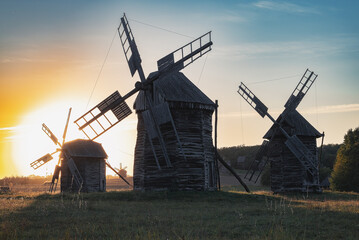 Naklejka premium Old wooden windmills at sunset in Pyrohiv open-air museum near Kyiv, Ukraine. Traditional rural landscape and heritage architecture in warm evening light.