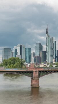 Aerial hyperlapse of Frankfurt skyline with Main River and Ignatz Bubis Bridge. Skyscrapers and church spires reflected in the water beneath dramatic clouds timelapse. Frankfurt, Germany