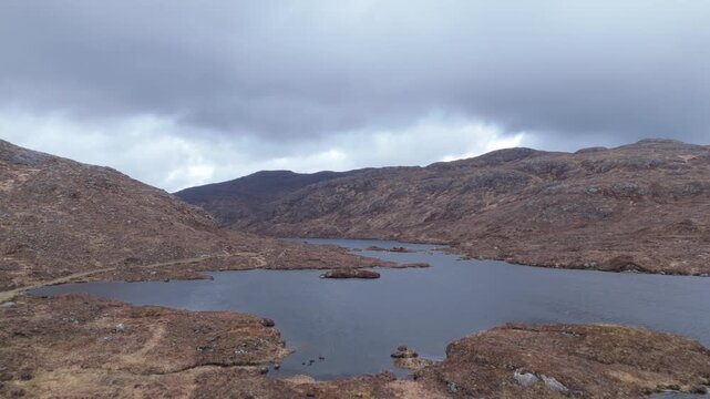 Drone glides backward from a wider perspective over an inland loch on the Isle of Harris, Scotland&mdash;revealing vast moorland, rocky hills, and a winding path under moody grey skies.