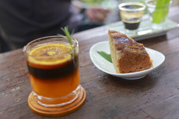 Refreshing iced coffee drink mixed with orange juice and slice of cake on wooden table. This relaxing beverage in glass with citrus garnish makes perfect cafe break