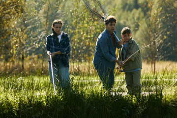 Caucasian middle aged man fishing with Caucasian teenage girl and Caucasian boy near grassy riverbank, holding fishing rods and net, all focused on water, outdoor nature setting