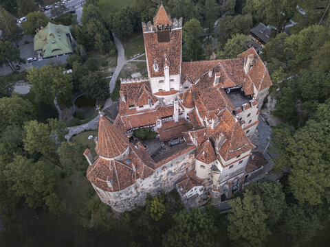 Aerial view of the imposing Bran Castle, its orange-tiled roofs and stone walls rising dramatically amidst a sea of deep green trees, Bucharest, Romania.